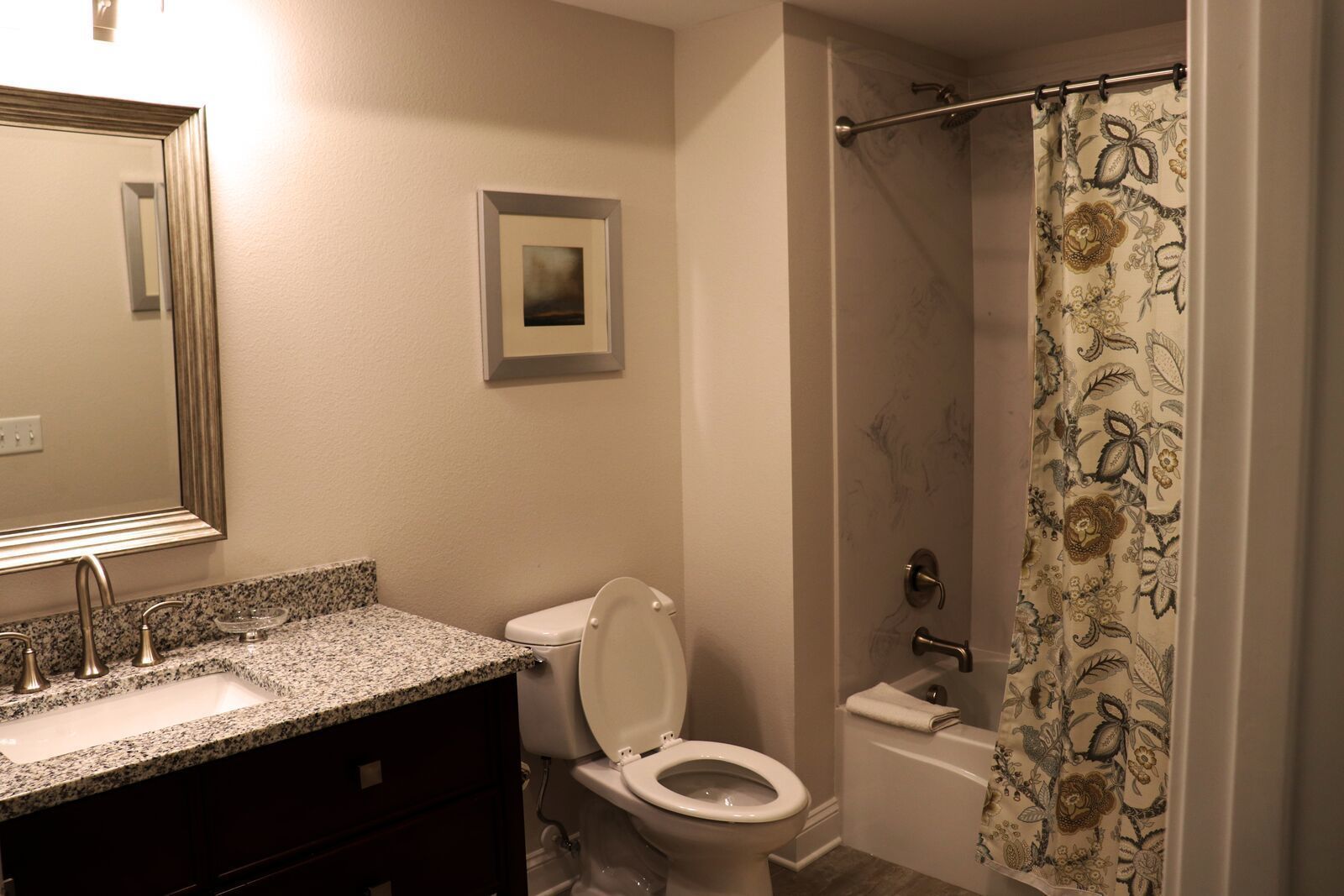 A bathroom with a granite countertop sink, a toilet, and a shower with a floral curtain.