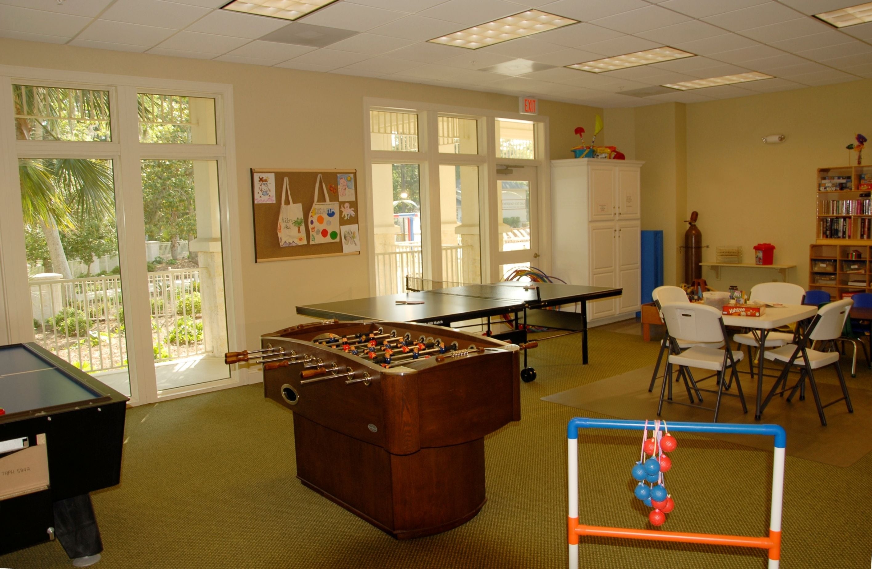 A room with a foosball table, ping pong table, and a foosball table in the corner. There is a window with a view of greenery outside.
