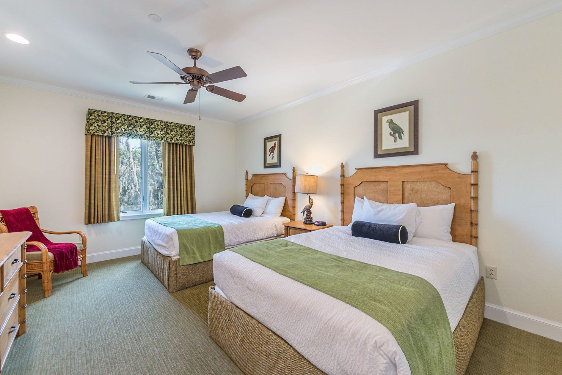 Une chambre d’hôtel avec deux lits simples, une tête de lit en bois, un bureau avec une chaise, une fenêtre avec des rideaux et un ventilateur de plafond.