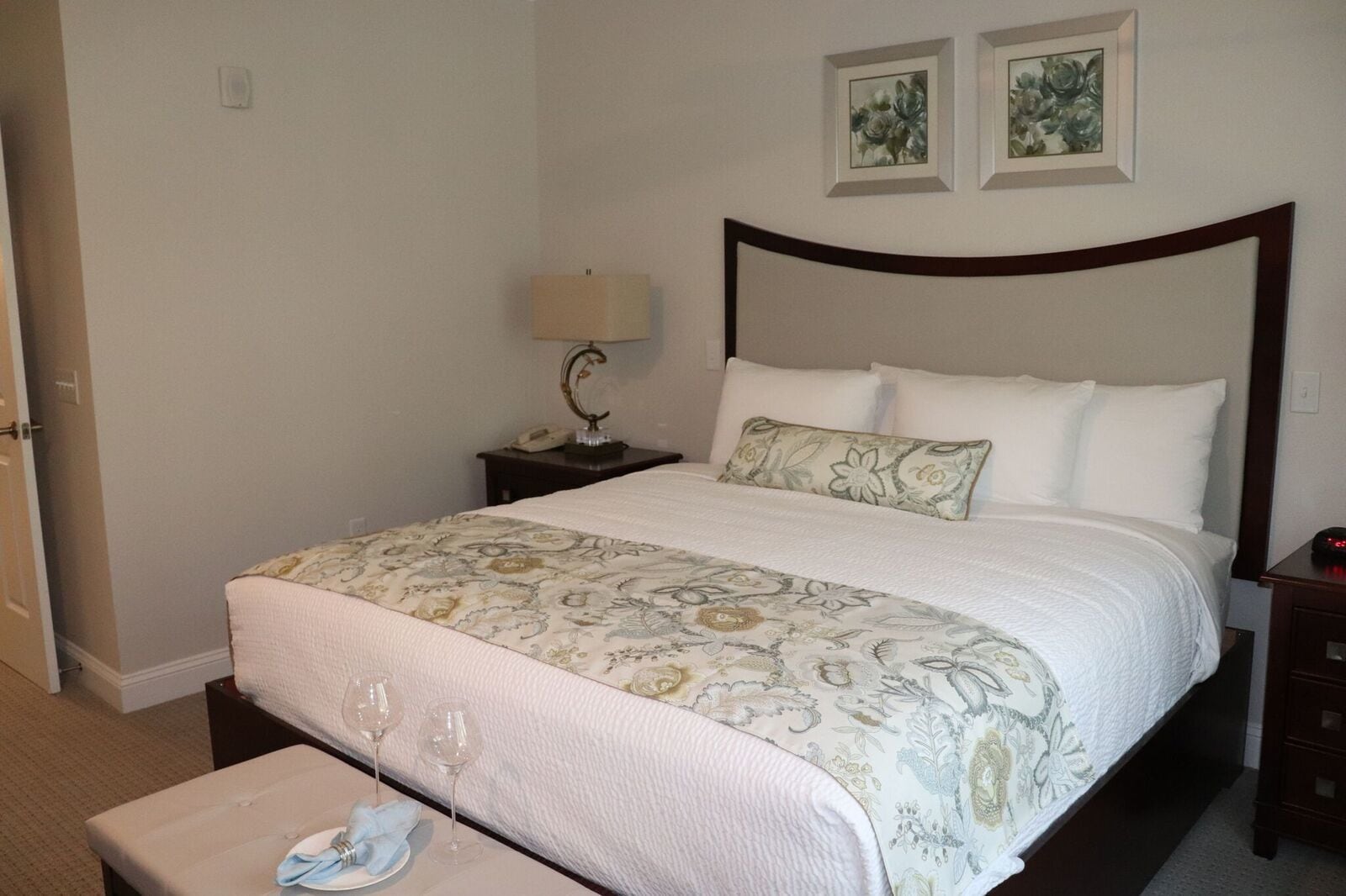 A neatly made bed with a floral bedspread, a bedside lamp, and two framed floral paintings on the wall.