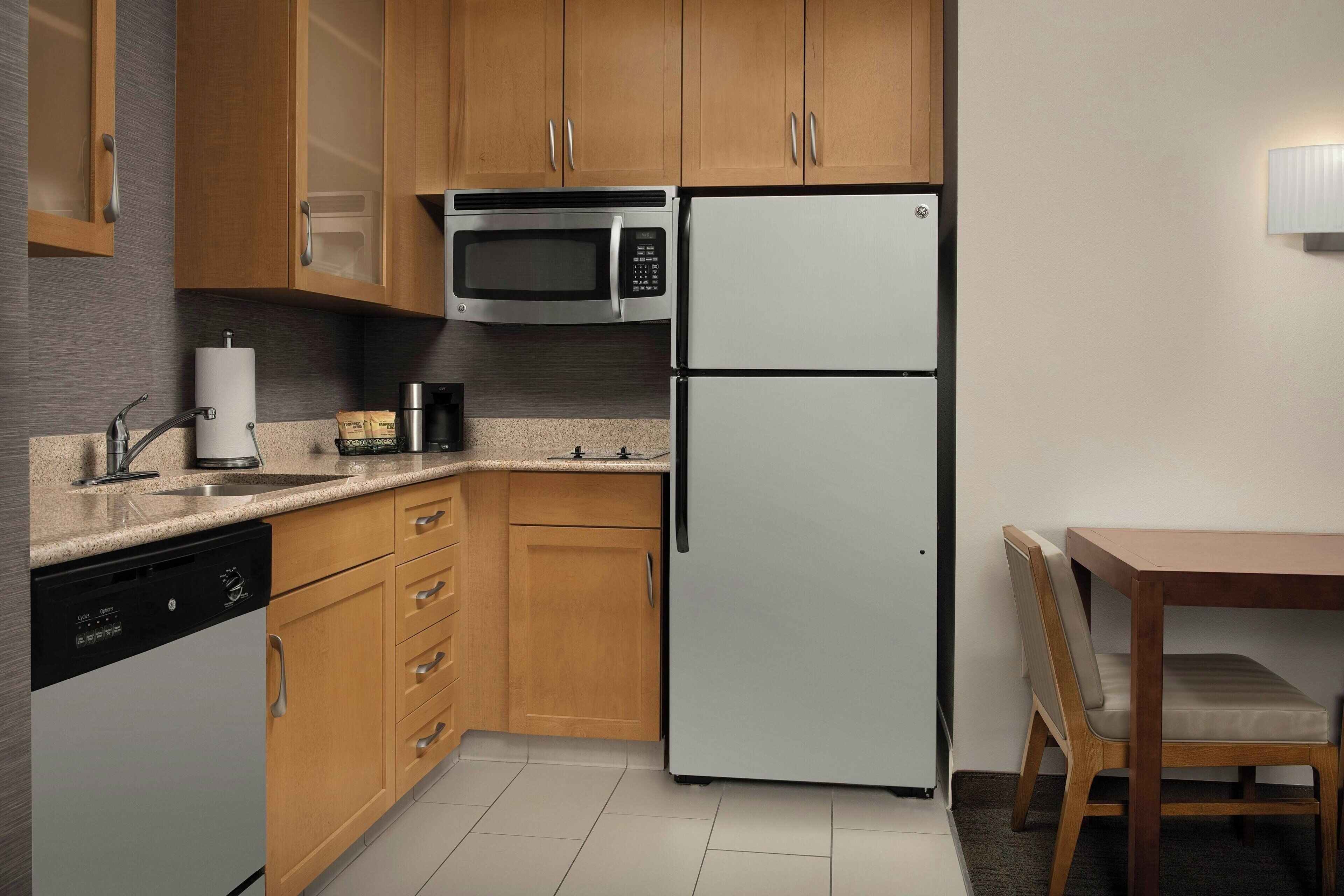 A compact kitchen with wooden cabinets, a microwave, a refrigerator, a dishwasher, and a dining table with a chair.