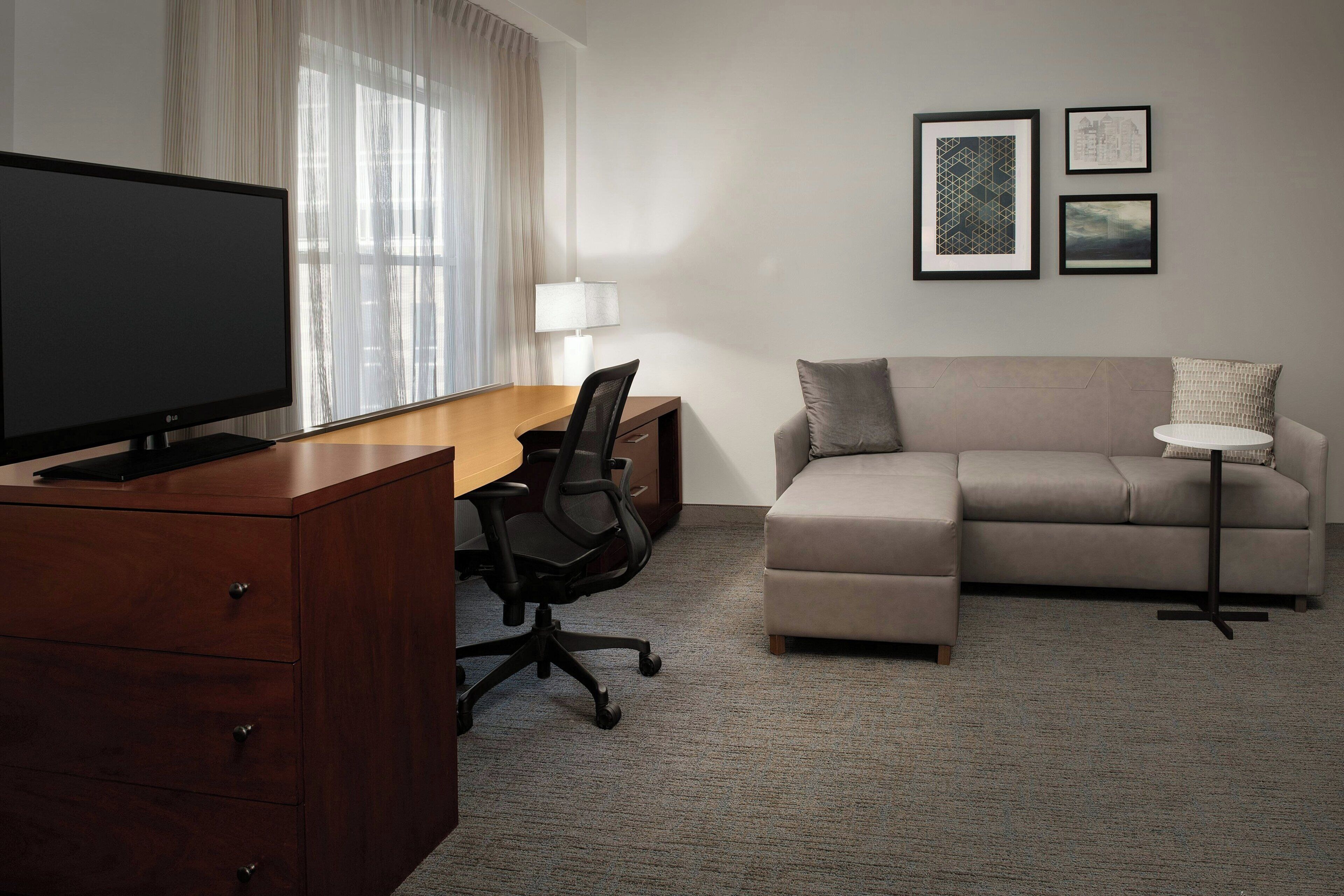 Een hotelkamer met een houten bureau, een flatscreen televisie, een leren bank, een klein rond tafeltje en twee ingelijste foto's aan de muur.