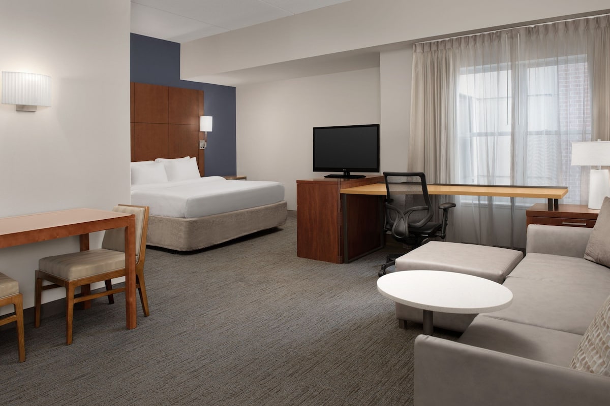 A hotel room with a bed, a desk with a computer, a chair, a sofa, a small table, and a window with curtains.