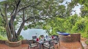 Een terras met een eettafel voor vier personen, omringd door rieten stoelen, met uitzicht op een meer en een kajak op een metselstenen rand.