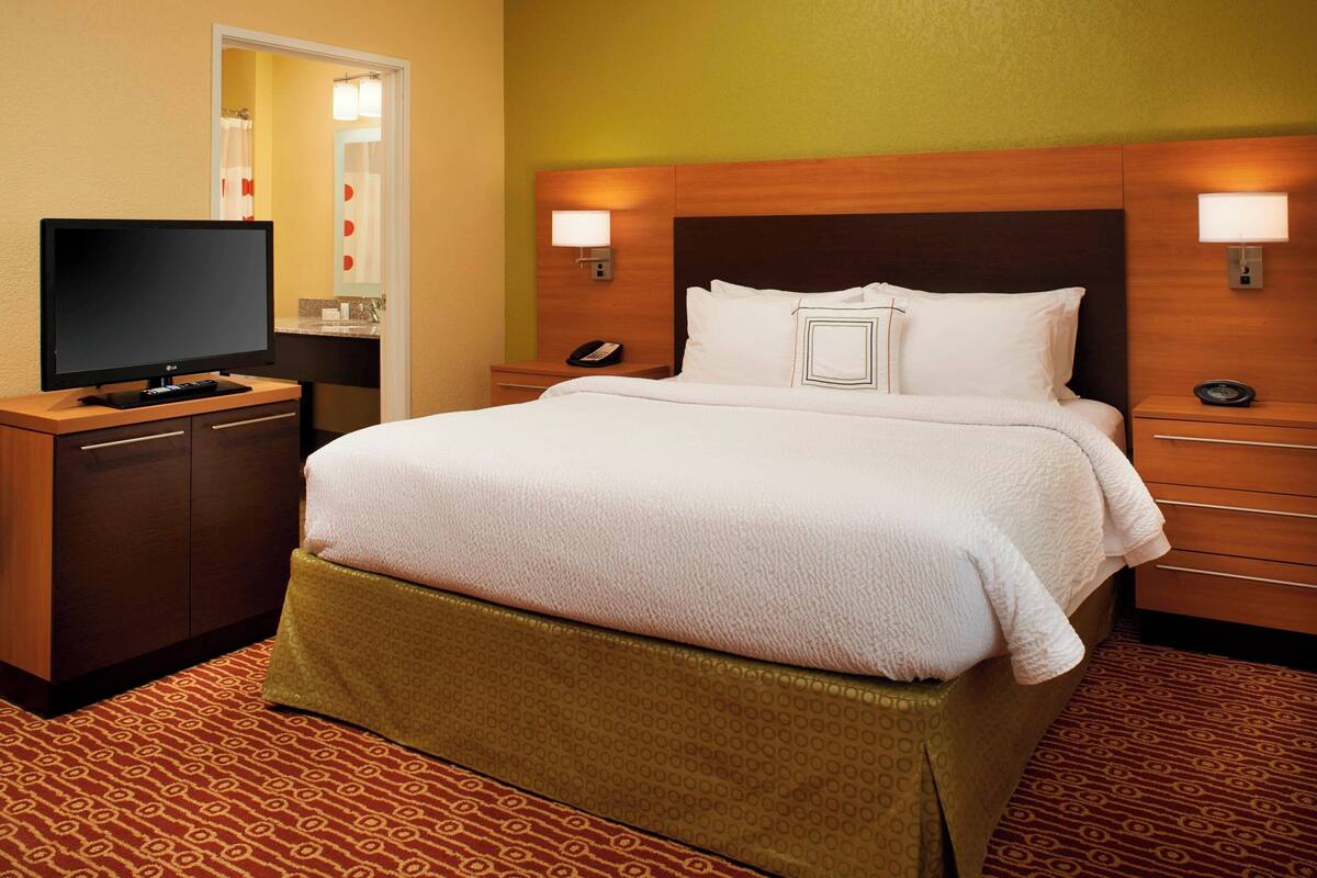 Habitación de hotel con una cama grande, un televisor sobre un mueble de madera y un baño visible a través de una puerta abierta.