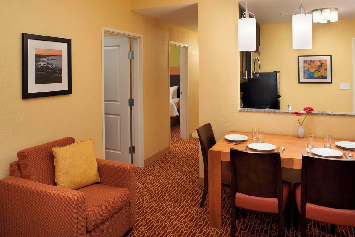 Habitación de hotel con zona de comedor, sofá con almohada, alfombra estampada y un cuadro en la pared.