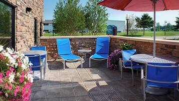An outdoor patio with blue chairs and a red umbrella, brick walls, and a view of a building in the background.