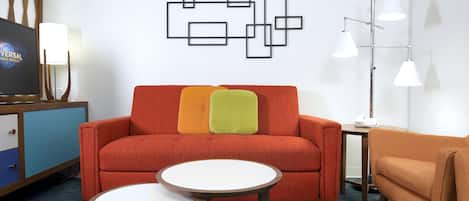 Una habitación de hotel moderna con un sofá, dos mesas redondas, un televisor y un adorno en la pared.