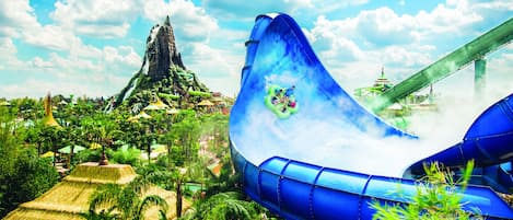 A water park with a large blue water slide, a mountain-like structure, and lush greenery.