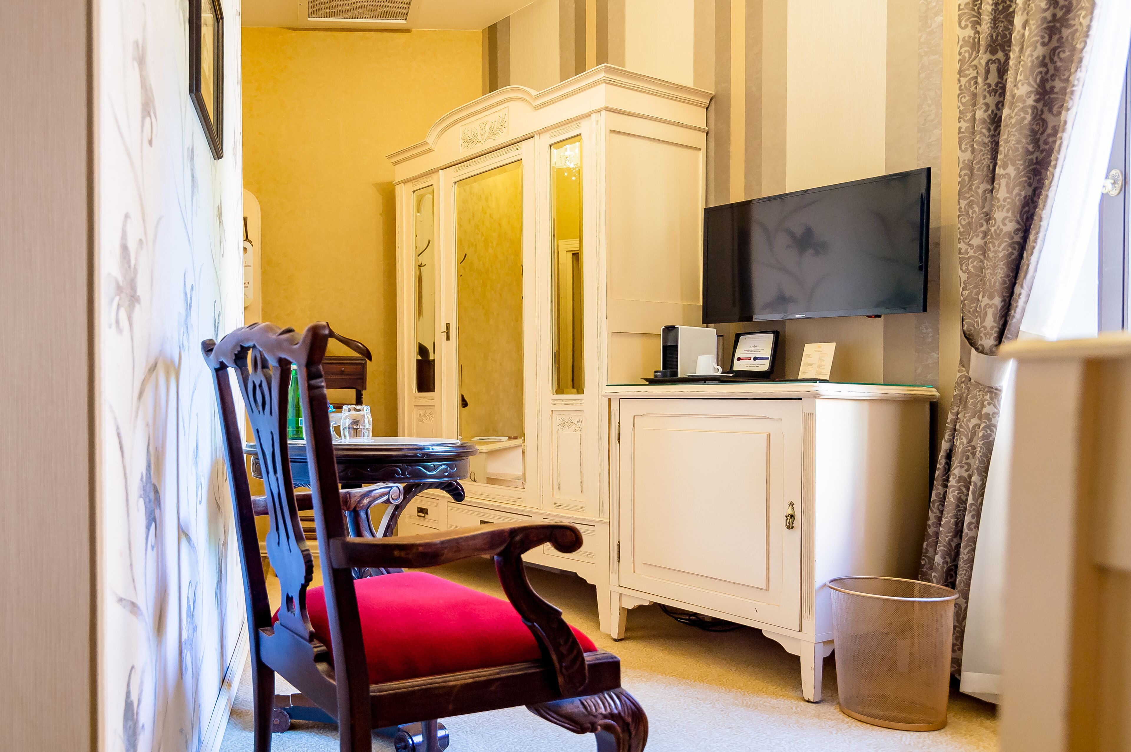 A hotel room with a wooden chair with a red cushion, a white cabinet, a television, and a trash bin.