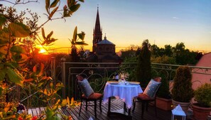 Un área de comedor al aire libre con una mesa y sillas, rodeada de plantas en macetas y vista a un edificio histórico con una torre de reloj.