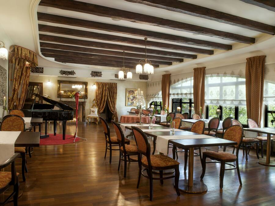 Un comedor con mesas y sillas de madera, un piano de cola y ventanas grandes con cortinas.