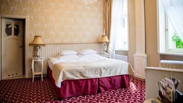 Quarto de hotel com cama, duas luminárias de cabeceira, radiador, janela com cortinas e tapete estampado.