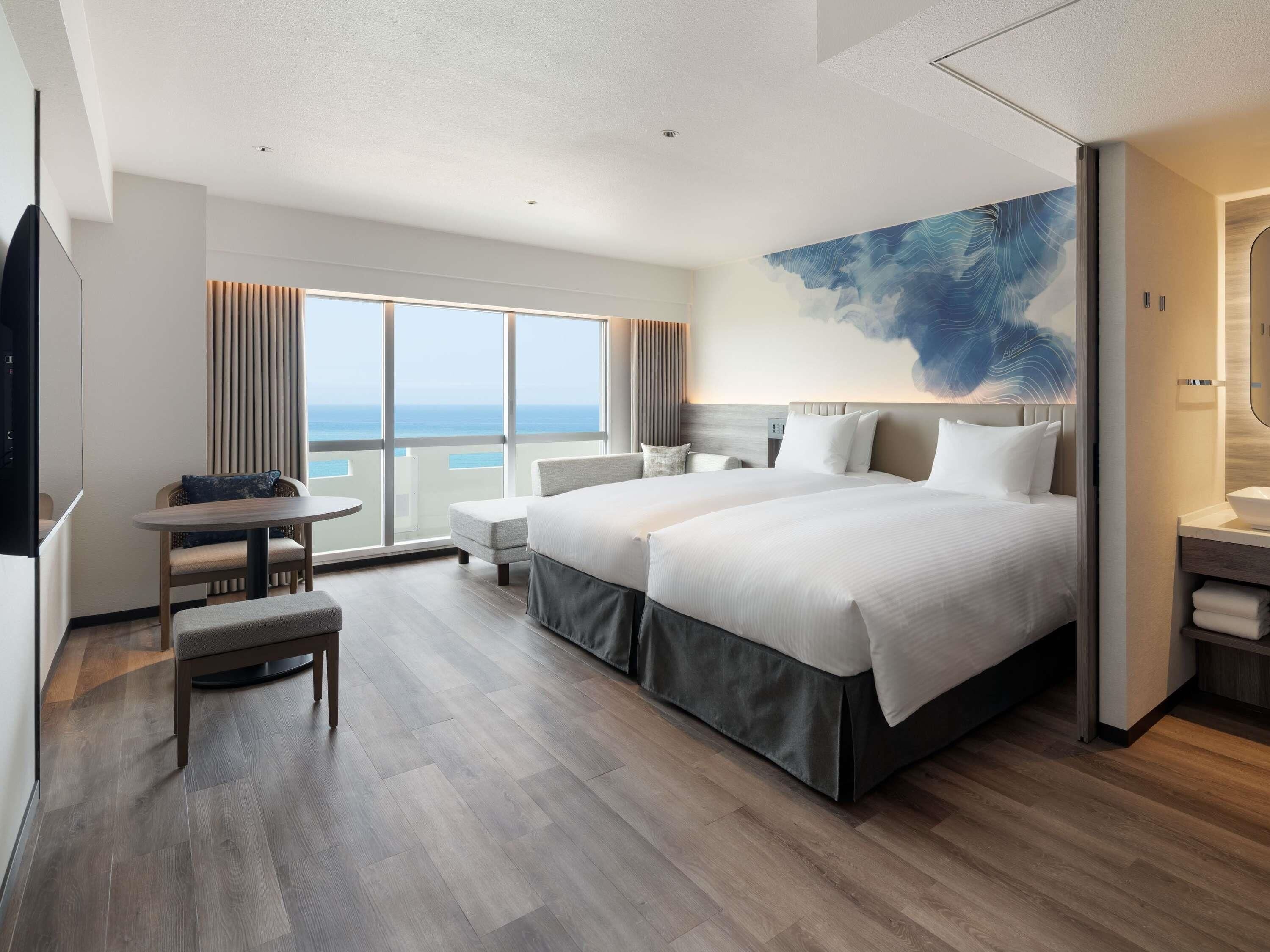 Quarto de hotel moderno com uma cama grande, uma secretária pequena, uma cadeira e vista para o oceano.