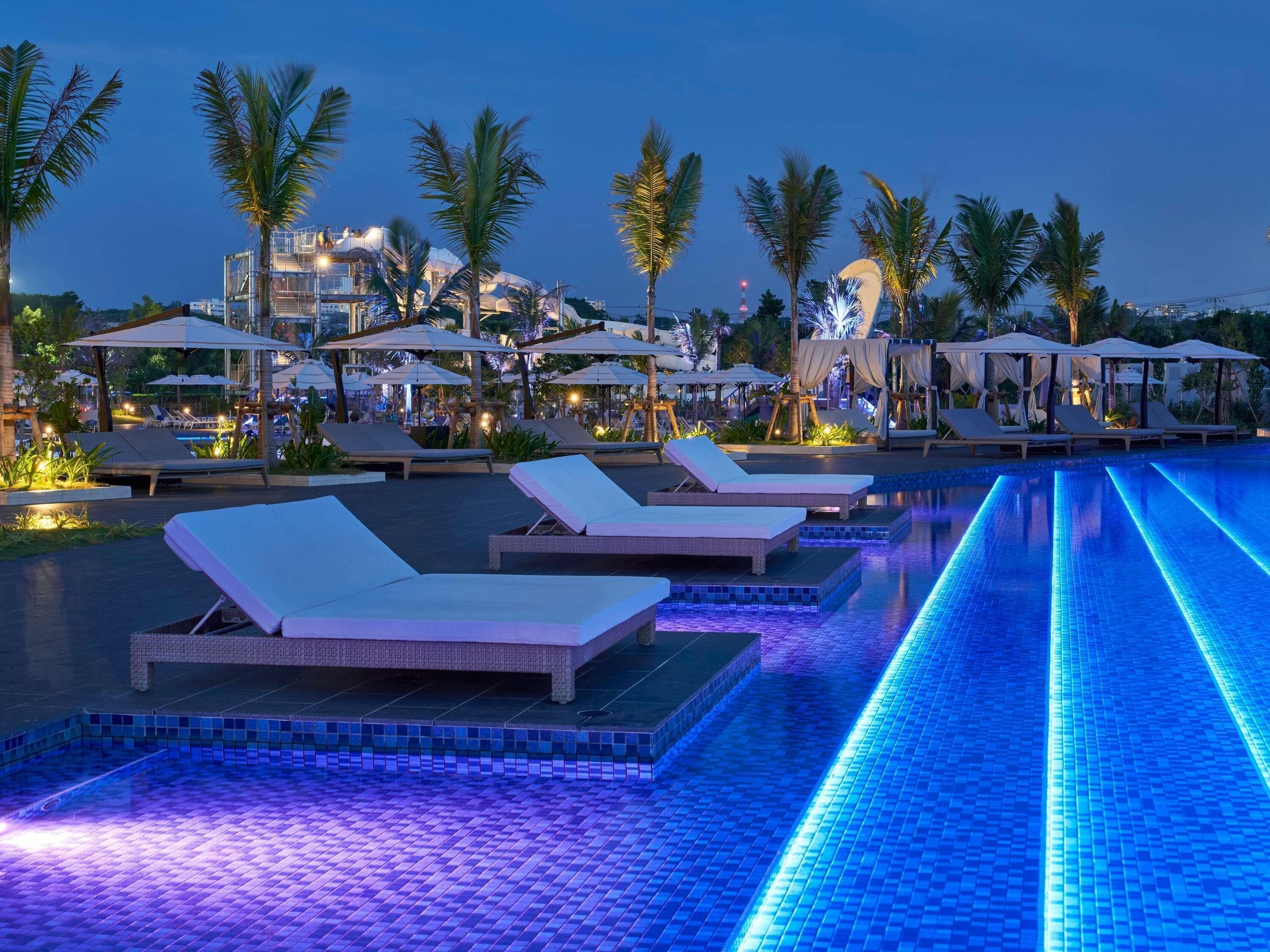 Un área de piscina con sillones de descanso y sombrillas, rodeada de palmeras.