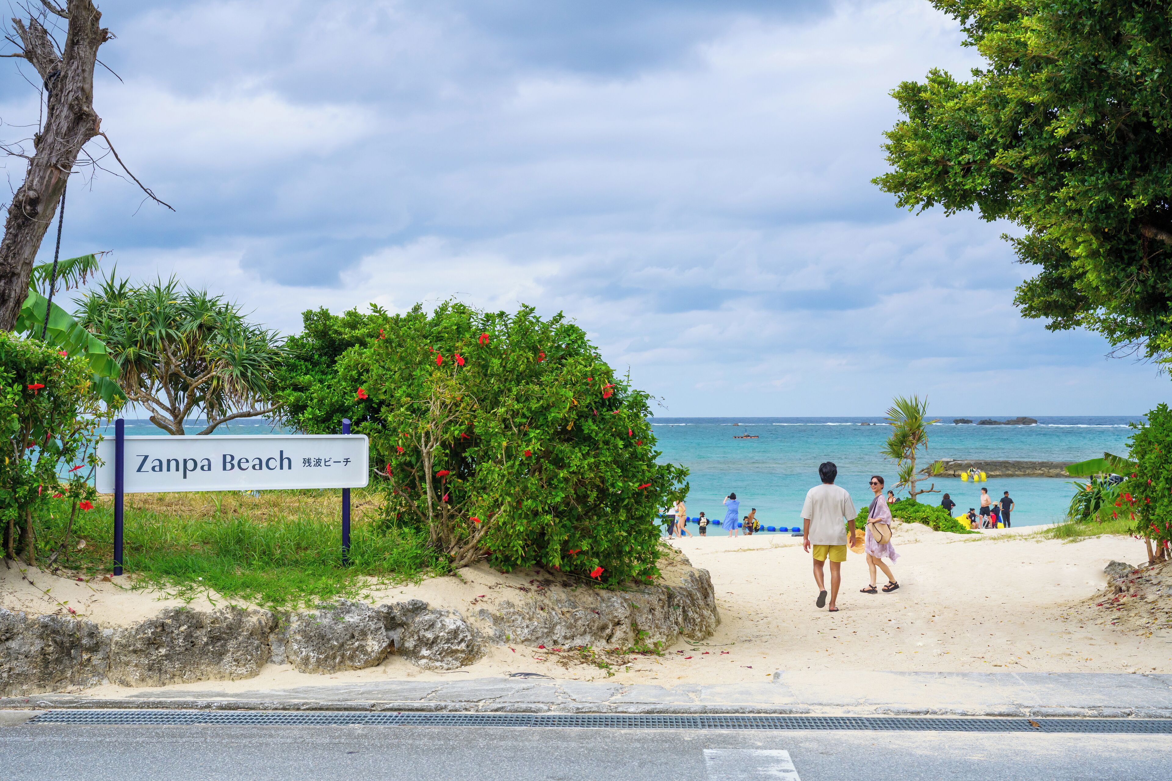 A beach sign for Zanpa Beach with a view of the ocean and people walking on the beach.