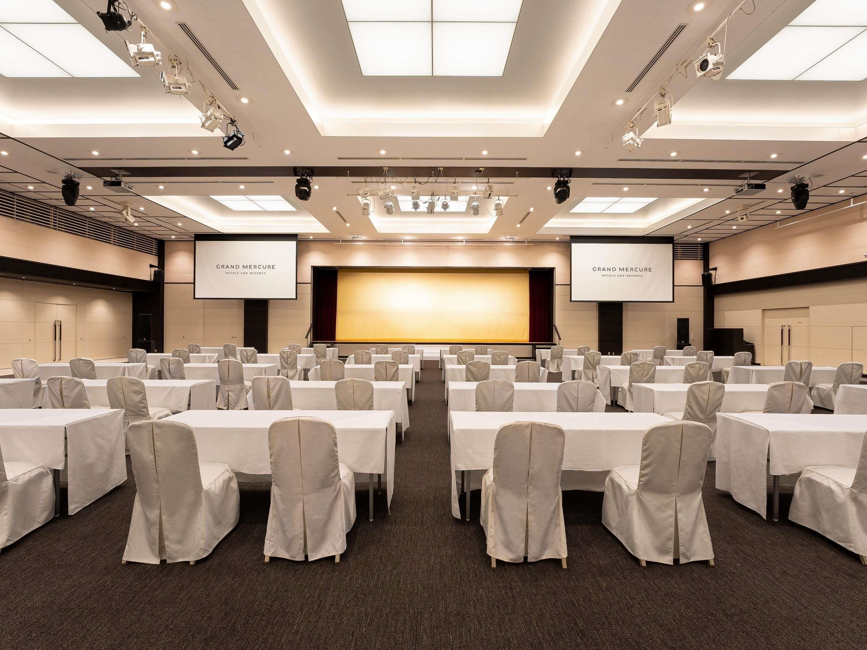 A conference room with rows of tables and chairs, a large screen, and a projector.