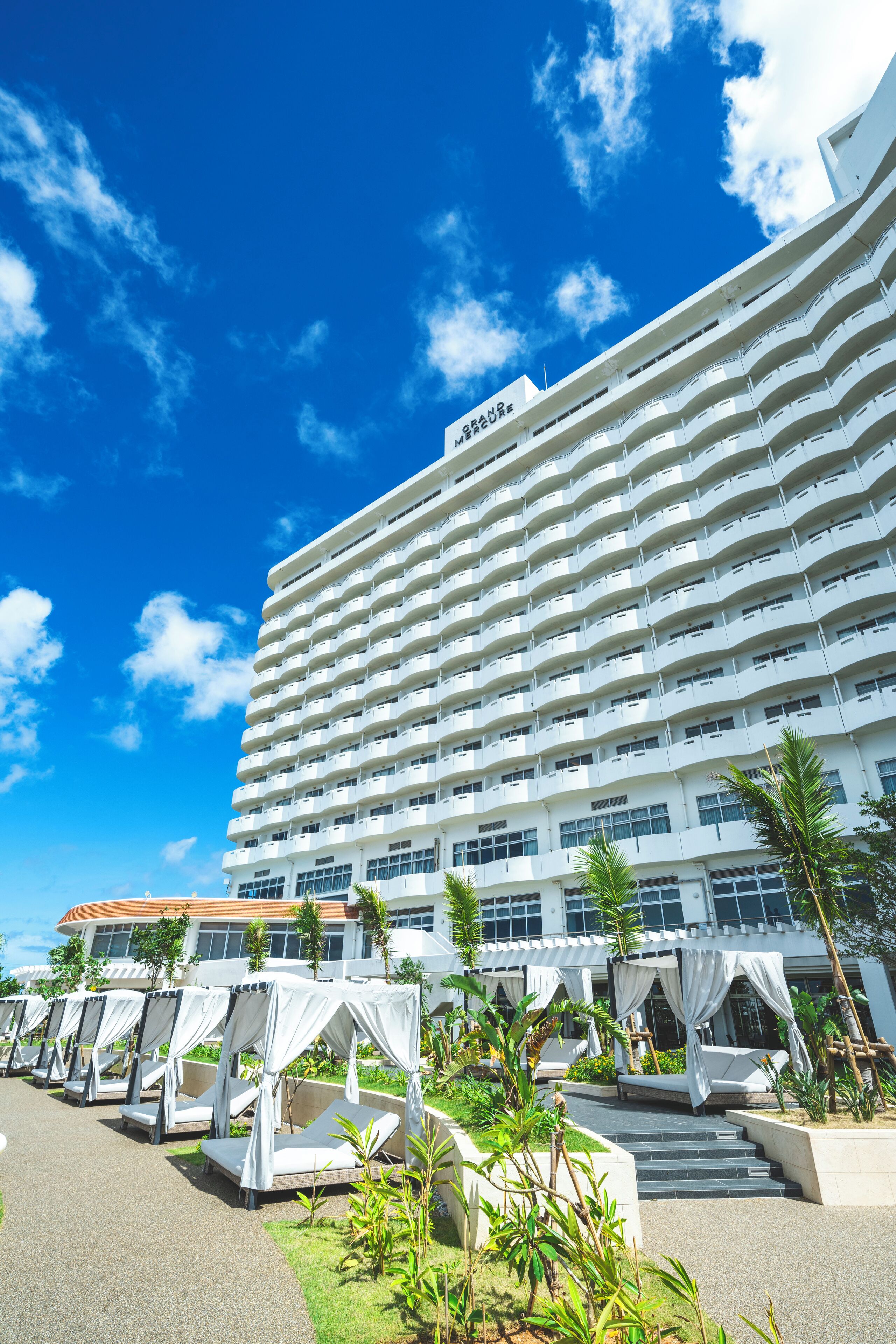 A multi-story hotel with a white facade, outdoor seating areas with white canopies, and a clear blue sky.
