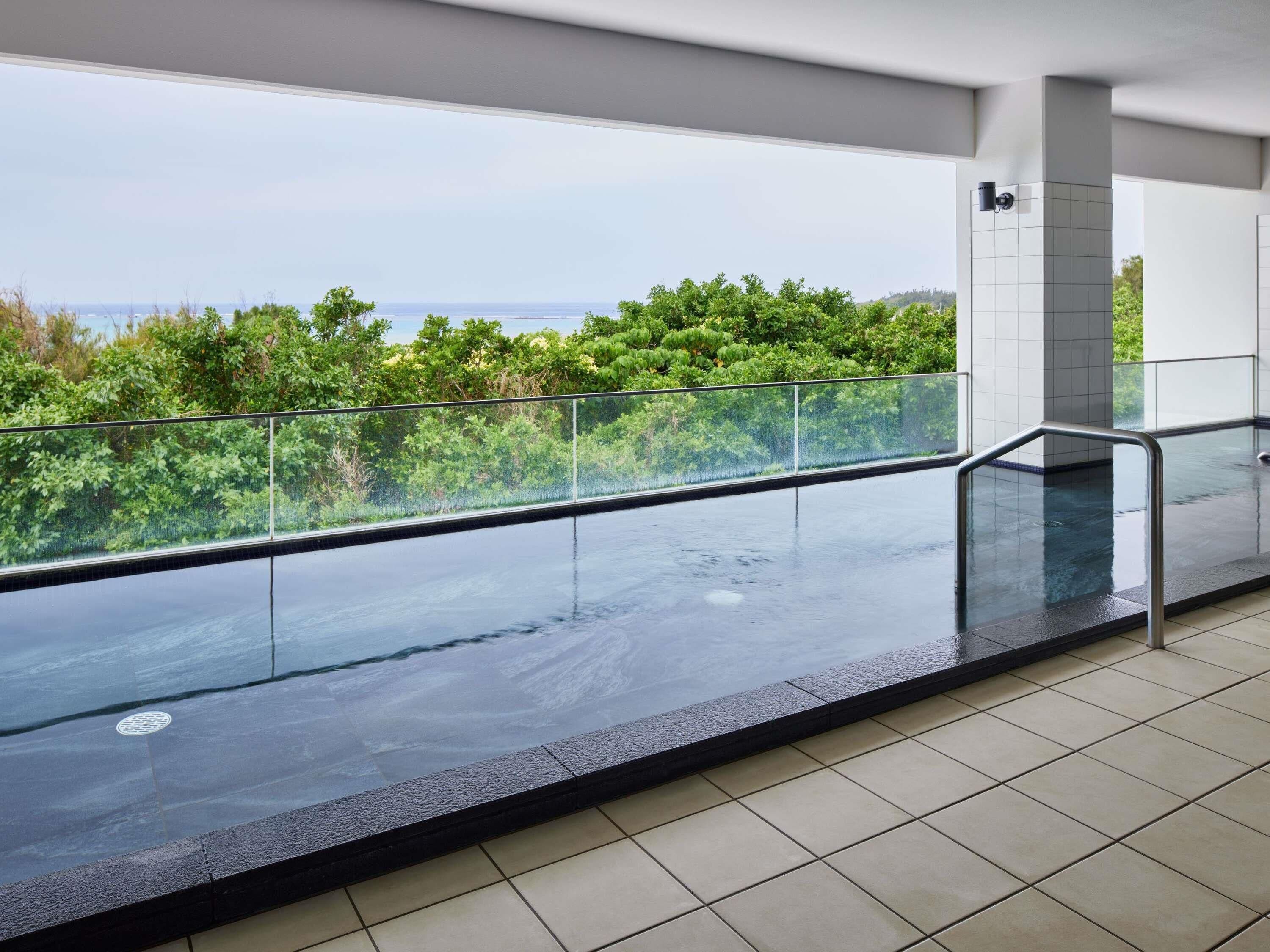 A modern indoor swimming pool with a glass barrier, a view of lush greenery, and a tiled floor.