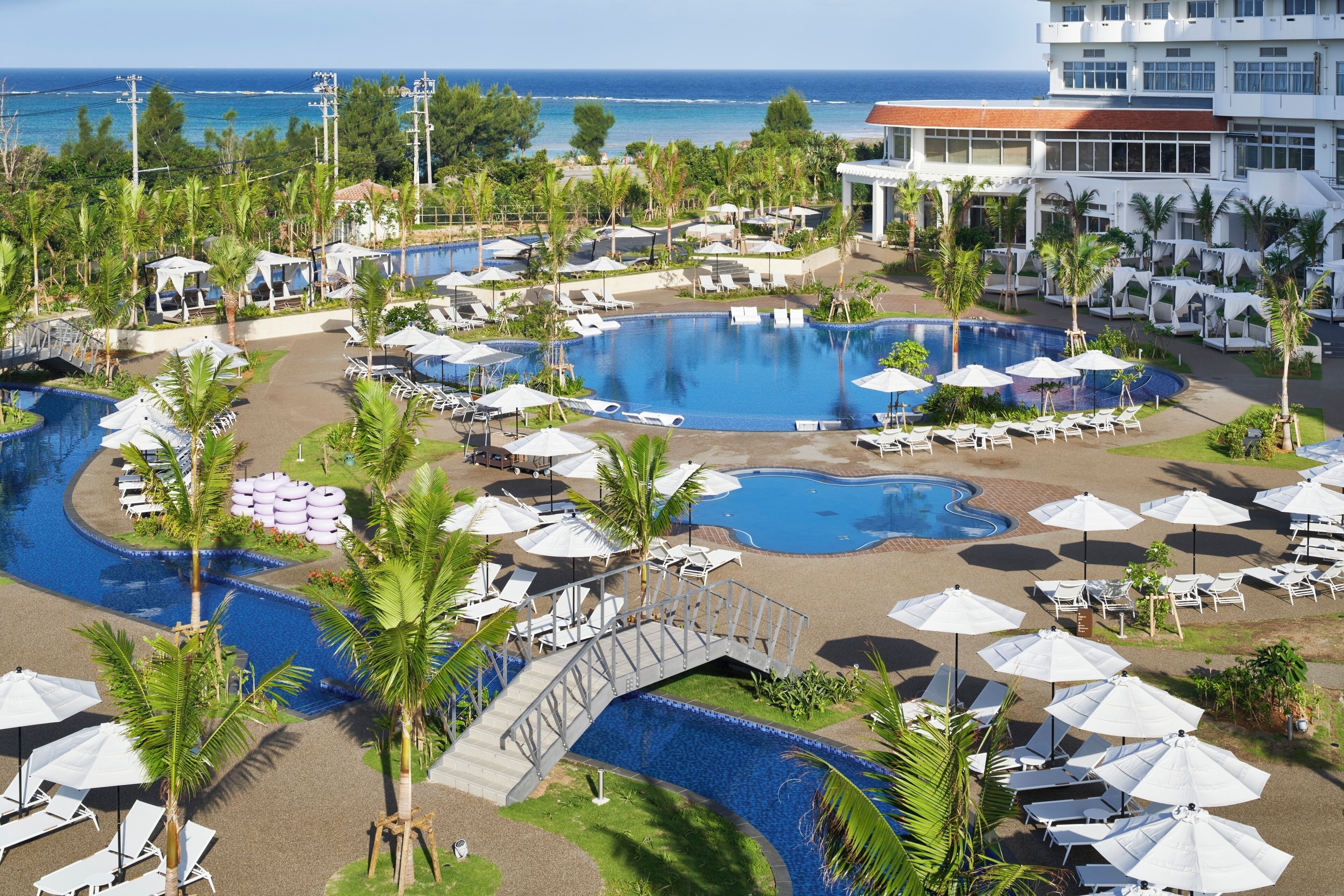 A resort with multiple swimming pools, sun loungers, umbrellas, and a bridge connecting different areas.