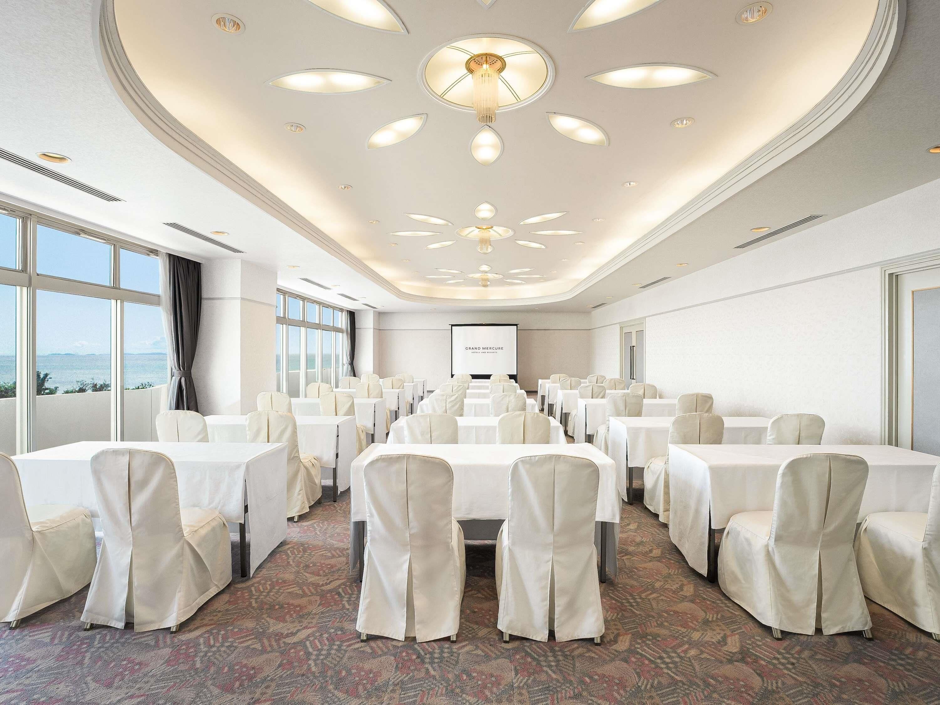 Ein Konferenzraum mit runden Tischen, weißen Stühlen, einer Projektionsleinwand und Blick auf das Meer.
