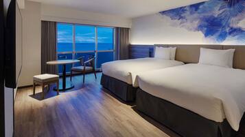 A hotel room with two beds, a small table, a chair, and a large window with a view of the ocean.