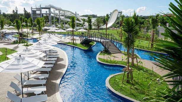 Un resort con una piscina central, varias zonas de descanso con sombrillas y un puente que conecta diferentes áreas.