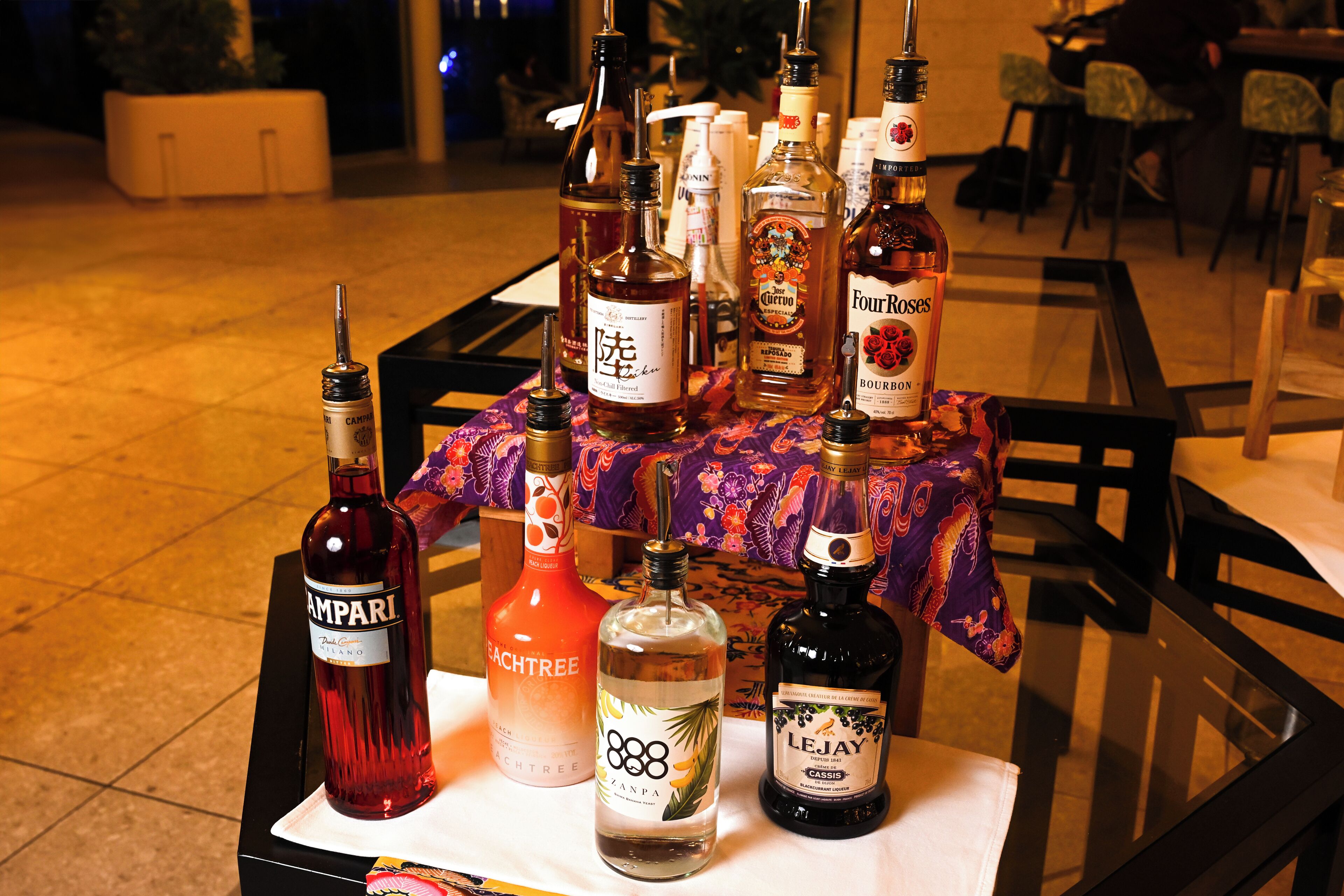 A table with various liquor bottles, including brands like Ampari, CactusTree, and Four Roses, set on a tablecloth with a floral pattern.