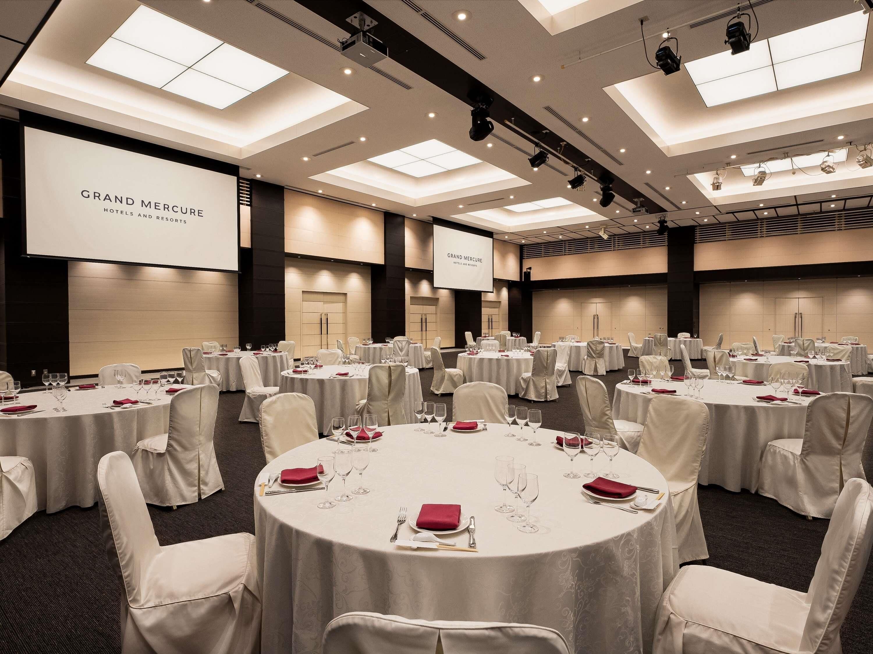 Un salón de conferencias con varias mesas redondas, sillas blancas y una pantalla grande que muestra 'GRAND MERCURE HOTELS AND RESORTS'.