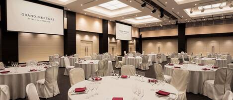 Une salle de conférence équipée de plusieurs tables rondes, de chaises blanches et d’un grand écran affichant le logo « GRAND MERCURE HOTELS AND RESORTS ».