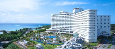 A large hotel with multiple floors, a pool area with water slides, and a view of the ocean.