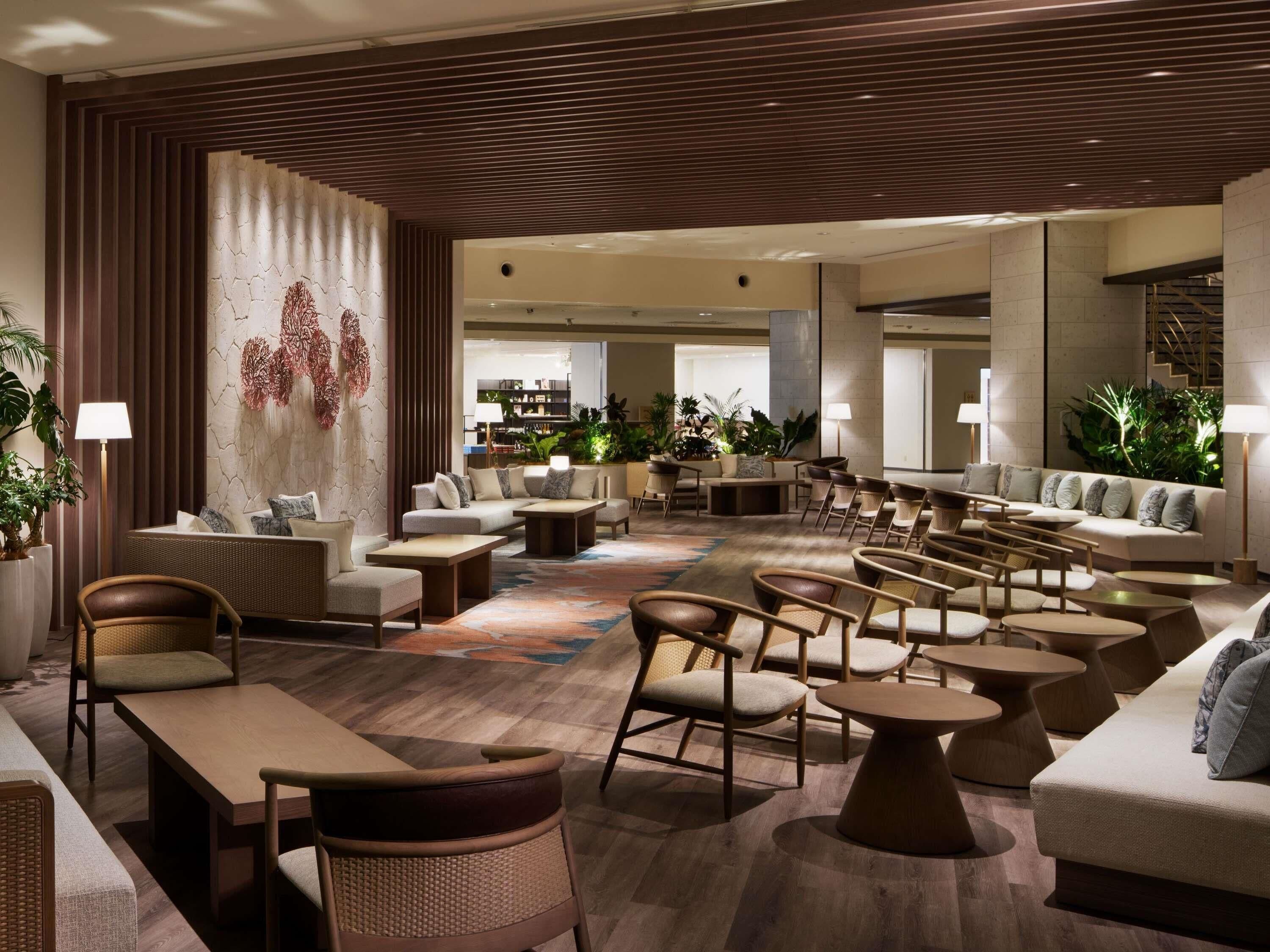 Een ruime lobby met diverse zithoeken, waaronder banken, stoelen en tafels. Er staan potplanten en een grote muur met een decoratief patroon.