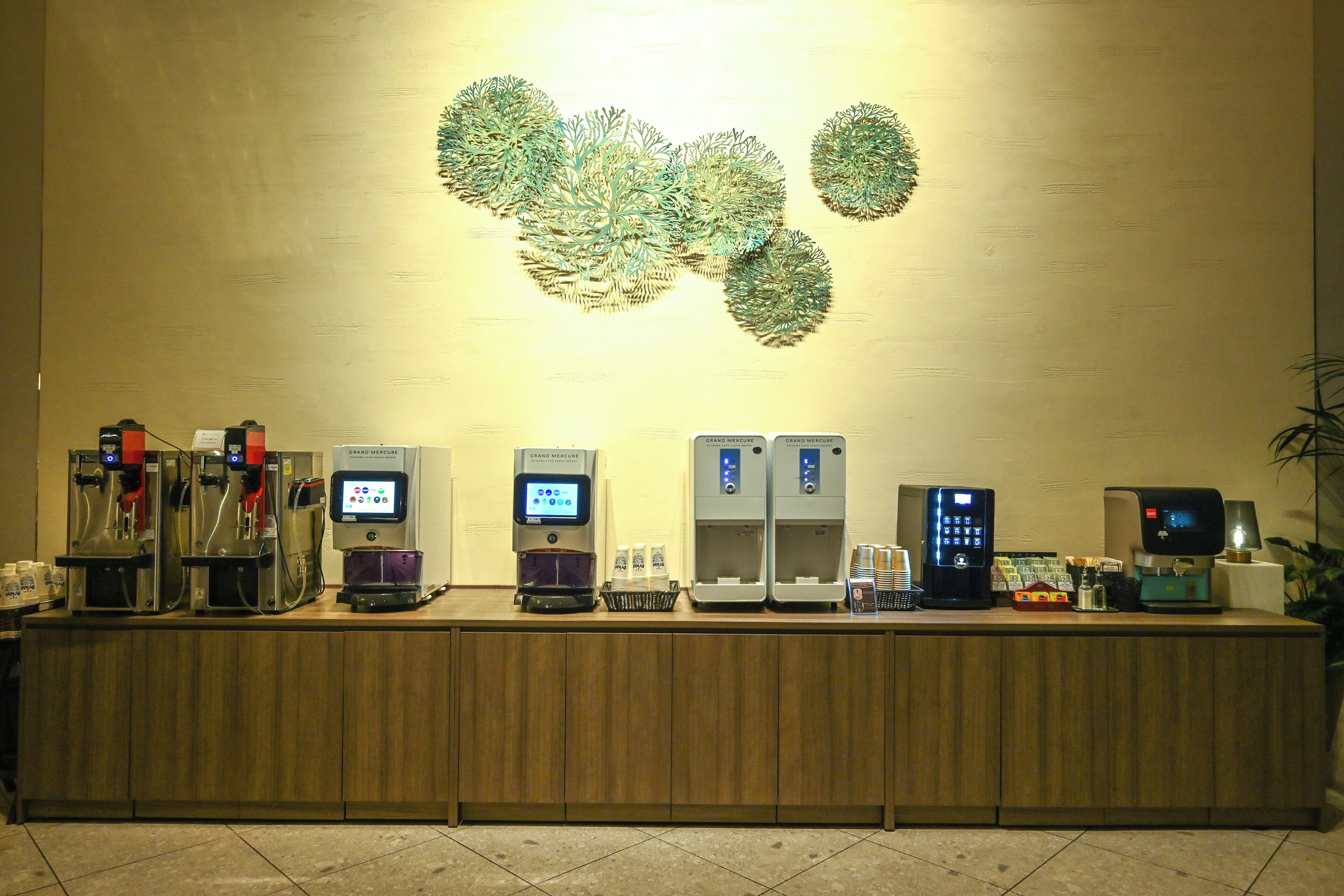 A hotel reception area with multiple coffee machines, a digital kiosk, and a decorative wall piece.