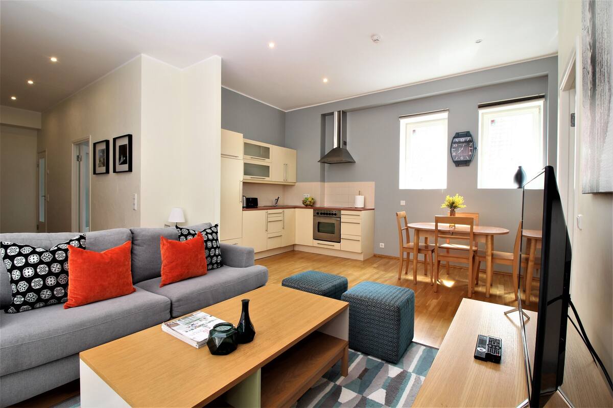 Un salon moderne avec un canapé gris, une table basse en bois et des poufs. On aperçoit, au fond, une cuisine avec des meubles blancs et une cuisinière.