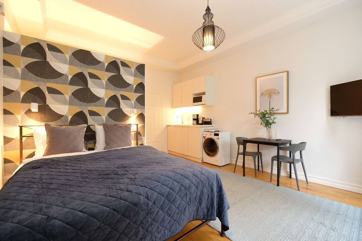 Une chambre moderne avec un grand lit, un lave-linge, un petit coin repas et un téléviseur fixé au mur.