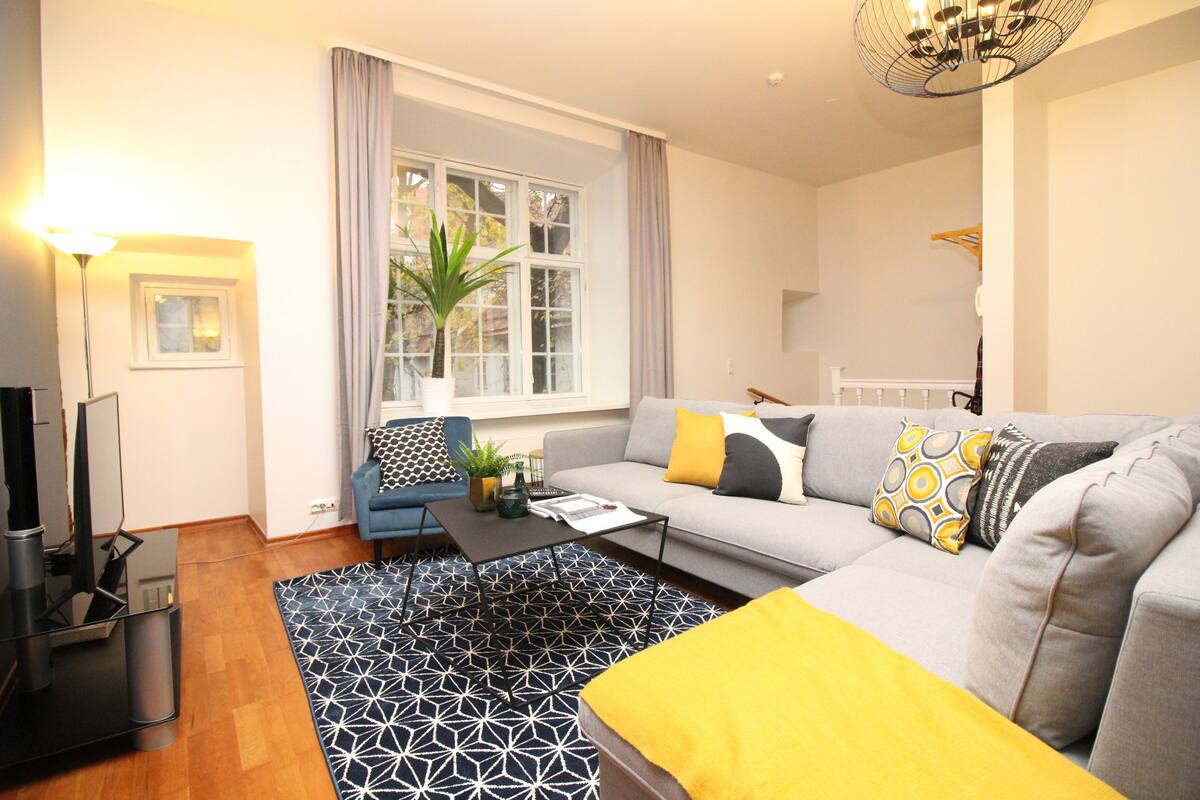 Un salon moderne avec un canapé gris, des coussins jaunes, un tapis à motifs noir et blanc et une grande fenêtre avec des rideaux.