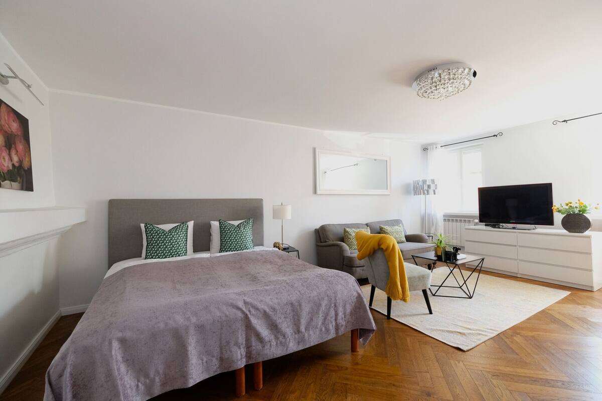 Une chambre à coucher moderne avec un grand lit, un fauteuil gris, une télévision et une commode blanche.