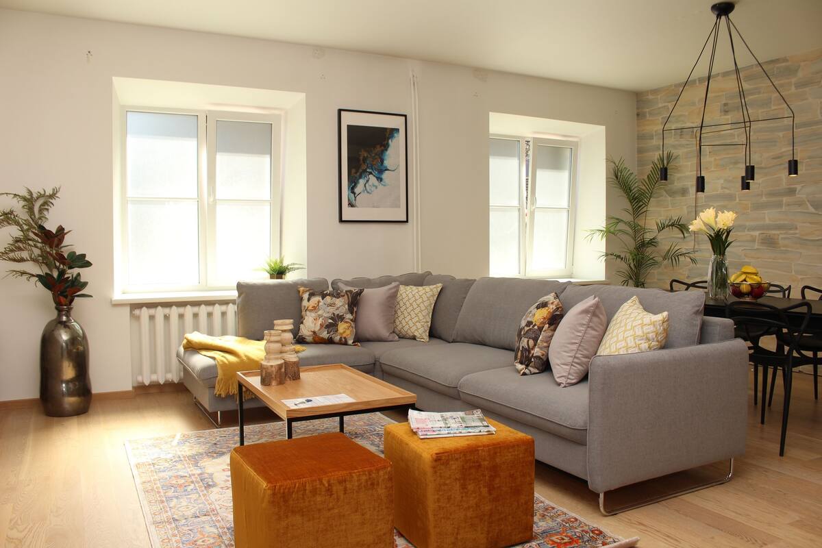 Un salon moderne avec un canapé gris, une table basse en bois et un pouf orange. Il y a deux fenêtres à cadres blancs, un radiateur et un mur en pierre agrémenté d’un luminaire suspendu.