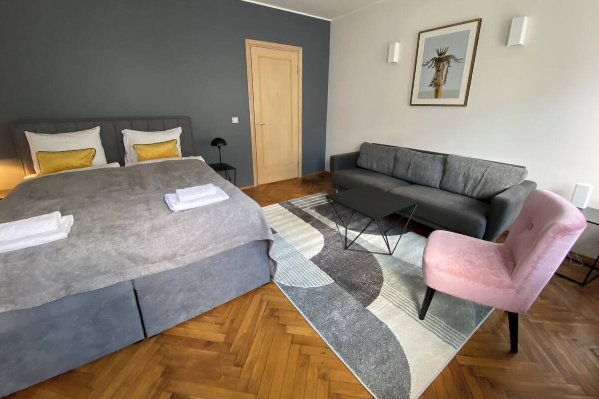 Une chambre à coucher moderne avec un grand lit, un canapé gris, un fauteuil rose et un plancher en bois.