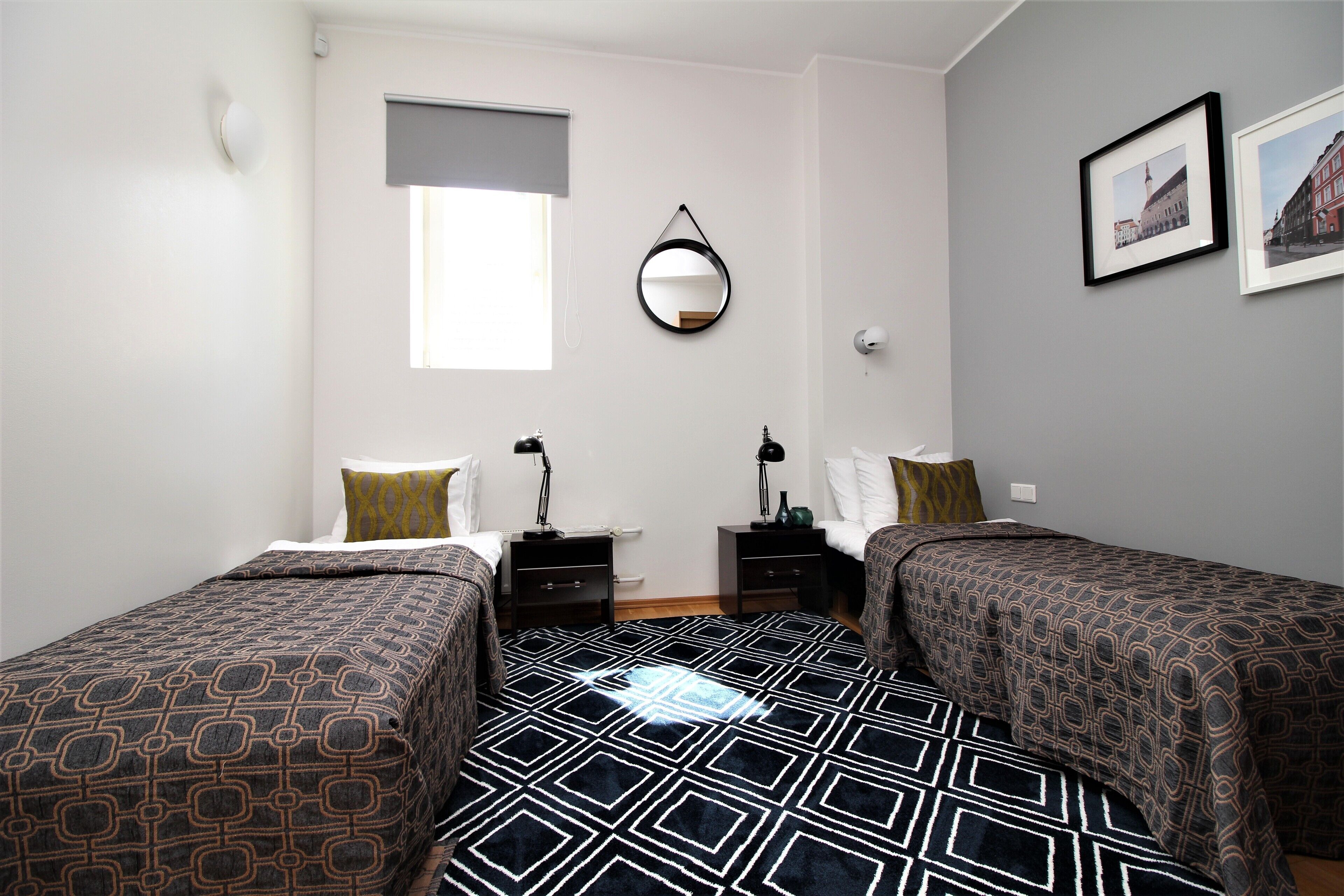 Slaapkamer met twee bedden, een raam met zonwering, een wandspiegel, nachtkastjes met lampen en ingelijste foto's aan de muur.