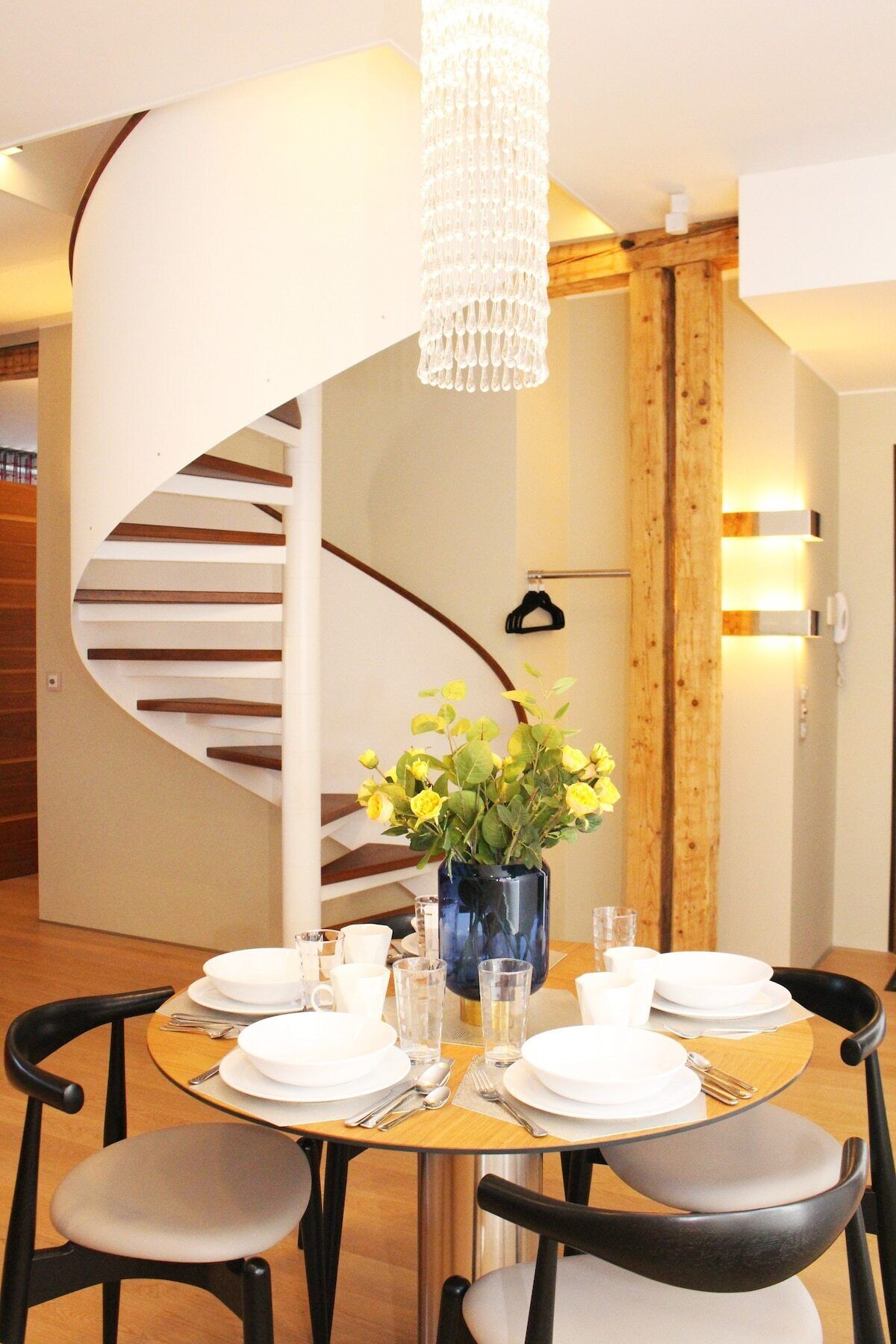 A dining area with a round table set for four, a vase with yellow flowers, and a spiral staircase in the background.