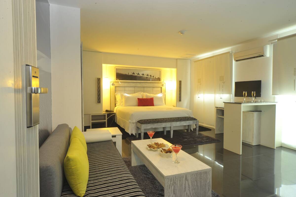 A modern hotel room with a bed, a sofa, a small dining table, and a kitchenette.