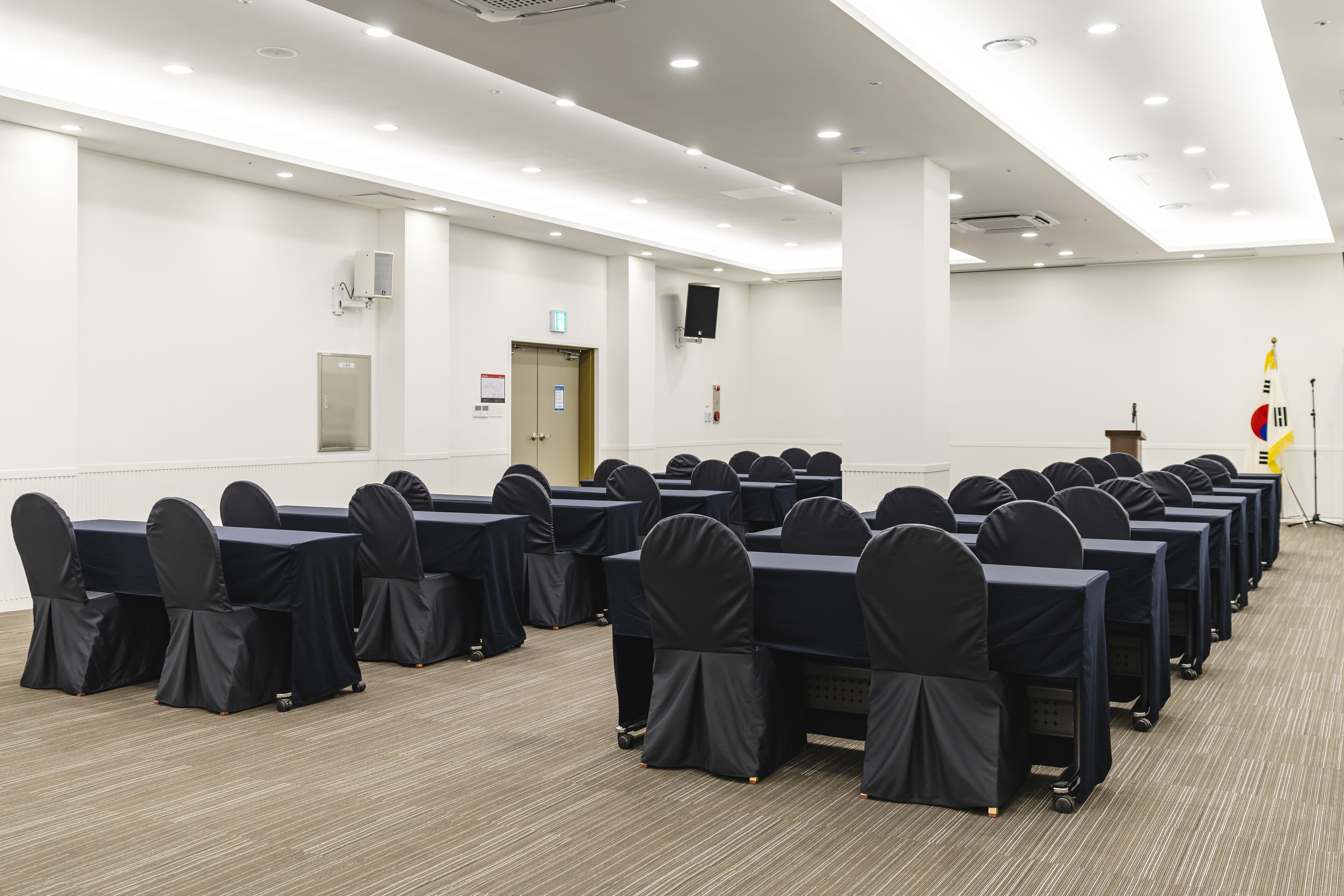 Un salón de conferencias con filas de mesas y sillas, equipado con luces en el techo y un atril.