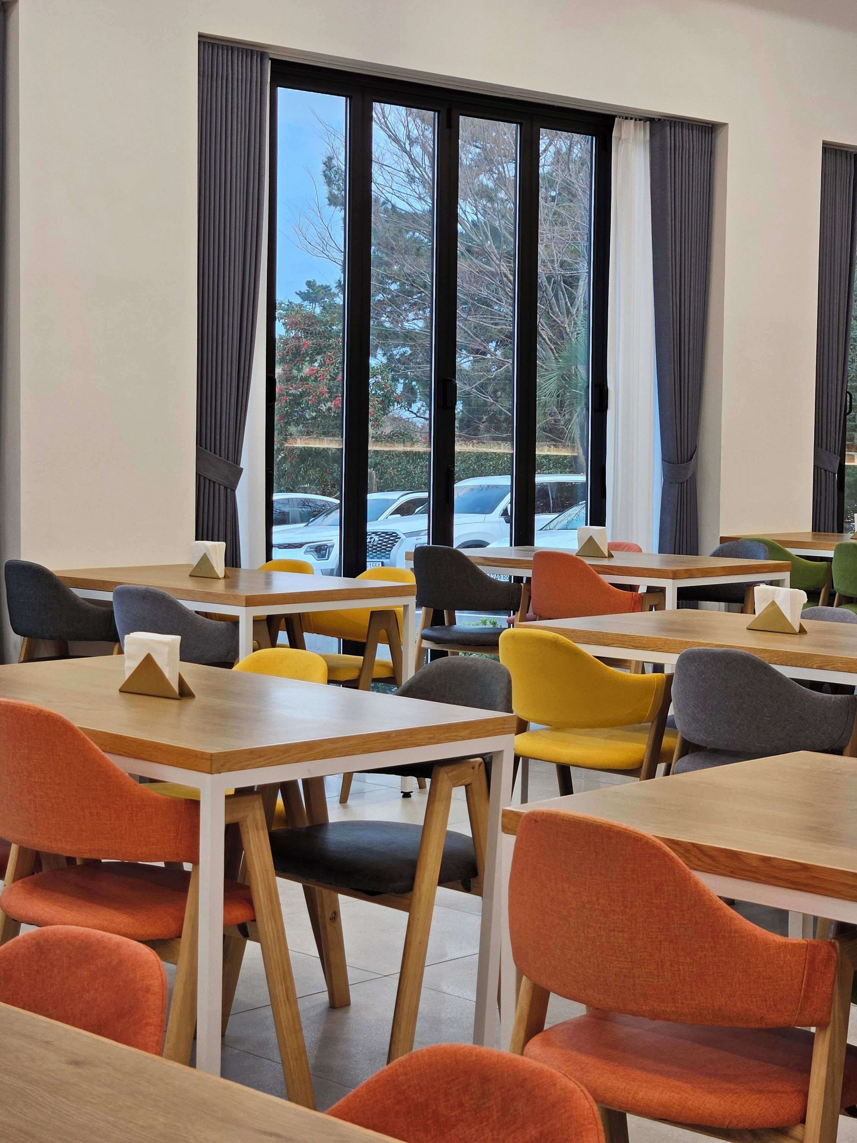 A modern dining area with wooden tables and chairs, large windows with curtains, and a view of parked cars outside.