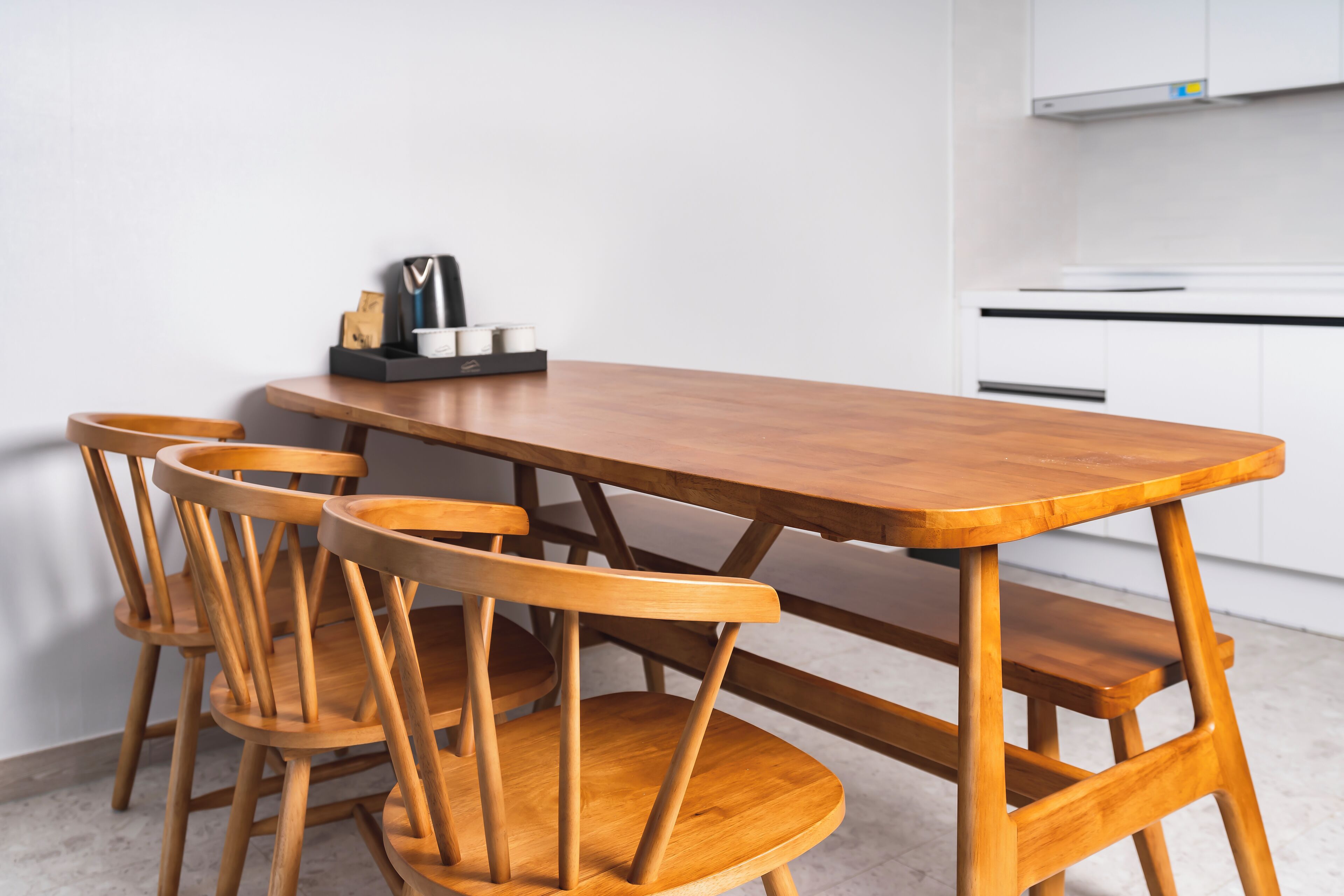 Una mesa de comedor de madera con cuatro sillas, una cafetera y una bandeja pequeña con tazas y una cuchara sobre una encimera blanca.