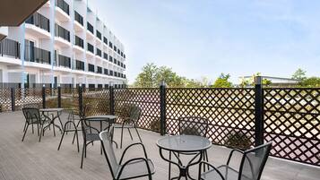 An outdoor patio area with tables and chairs, a wooden deck, and a building with balconies in the background.