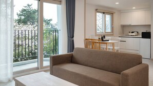 A modern living room with a sofa, a dining table with chairs, a kitchen area, and a balcony with a view of trees. - Hallim Resort (Jeju City)