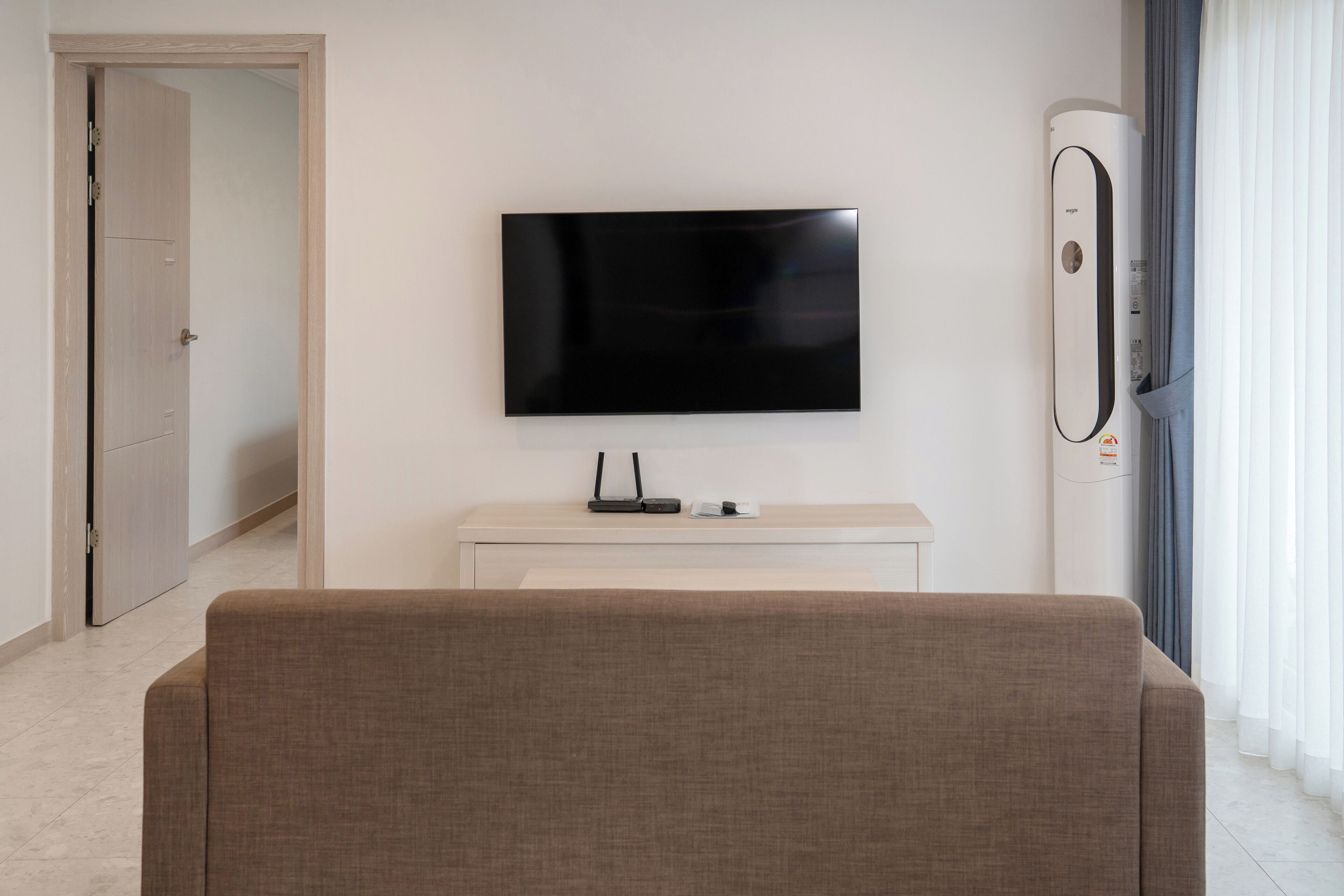 Ein modernes Wohnzimmer mit einem flachbildfernseher an der Wand, einem weißen Konsolentisch, einem braunen Sofa und einem großen Fenster mit Vorhängen.