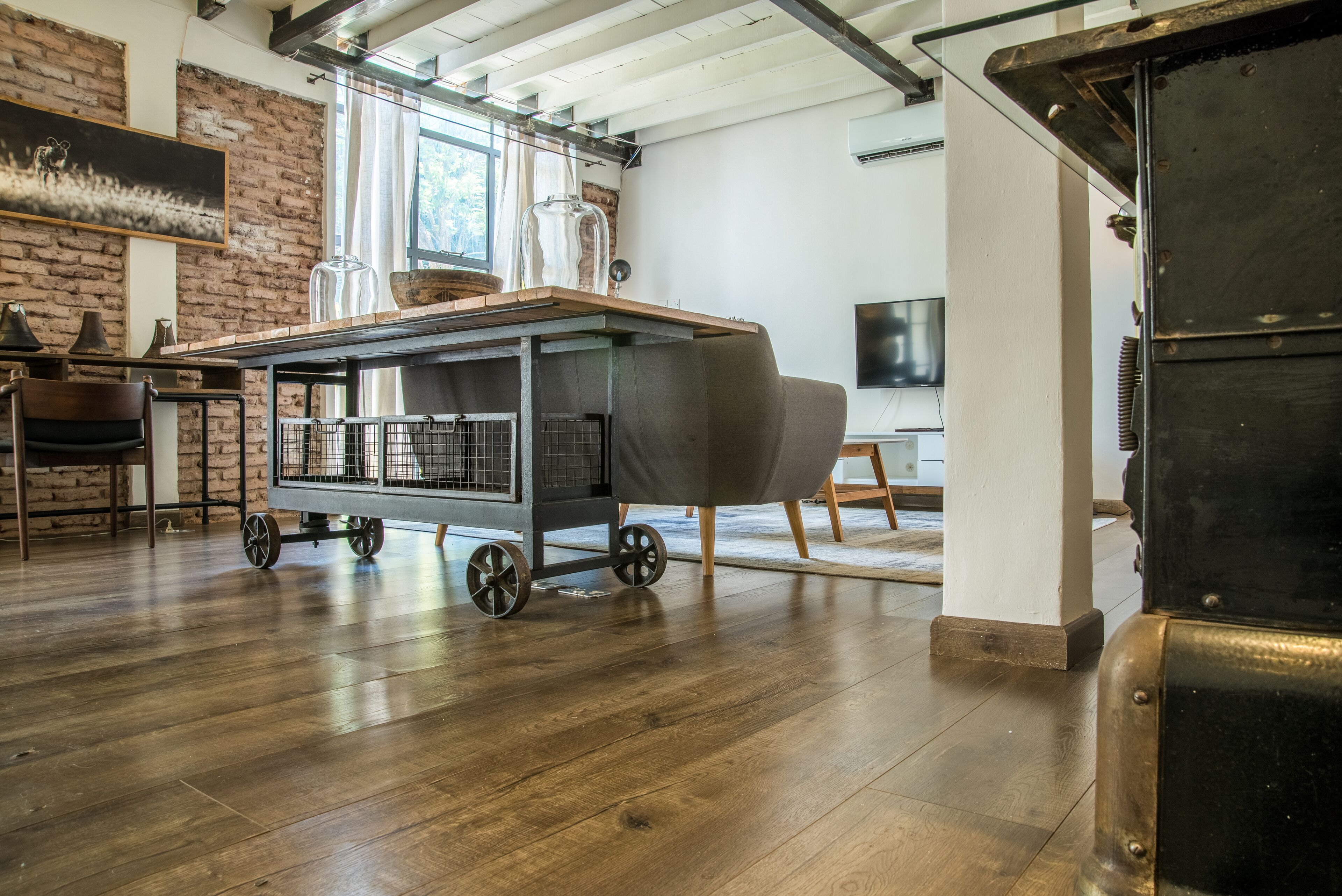 A spacious room with a wooden floor, a cart with a covered grill, a brick wall, and a large window.