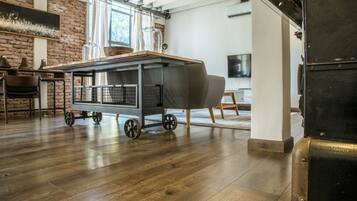 A spacious room with a wooden floor, a cart with a covered grill, a brick wall, and a large window.